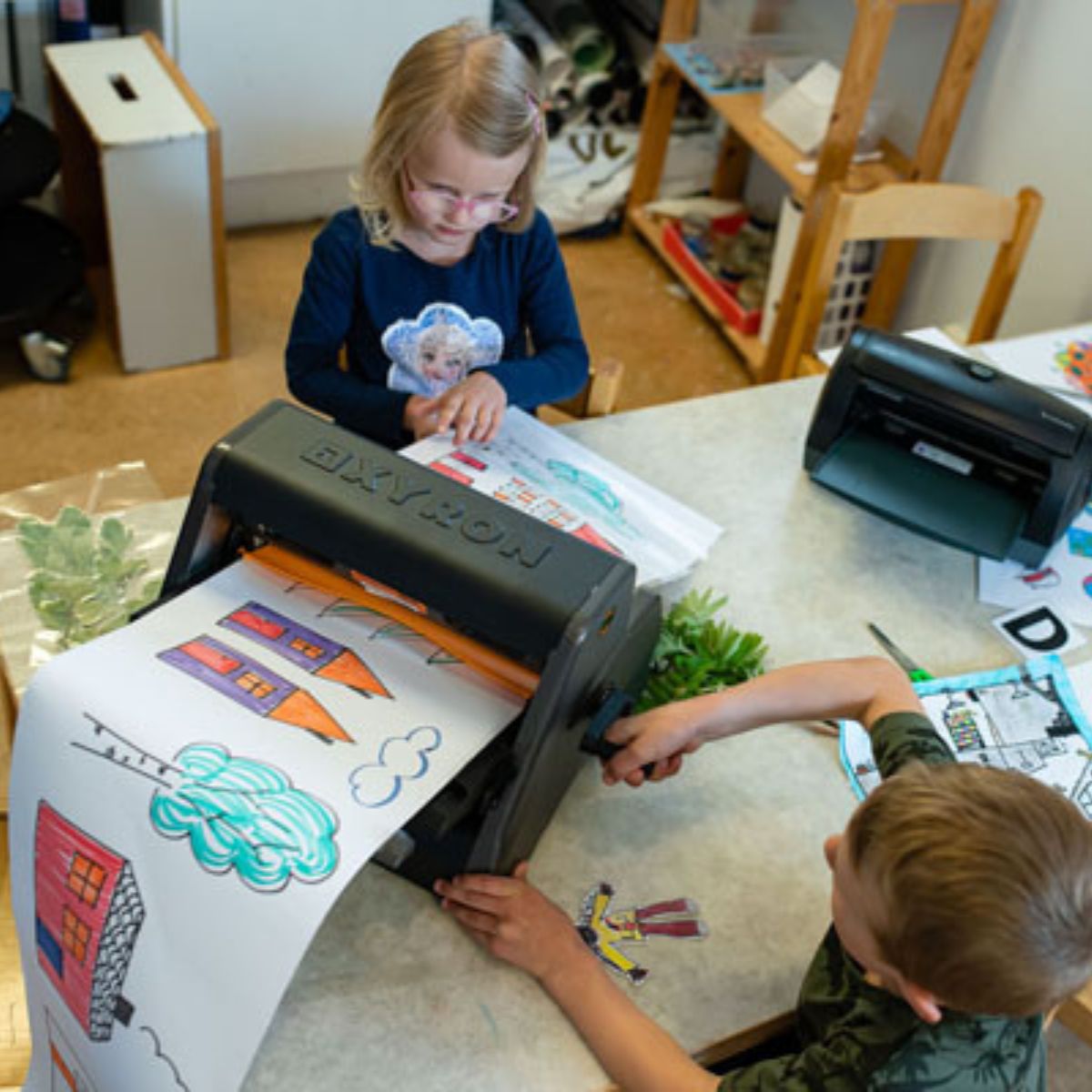 En flicka och en pojke använder en kallamineringsmaskin där de laminerar en avlång teckning som dem har målat.